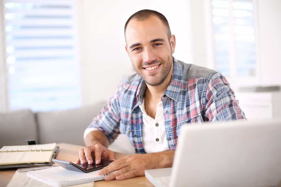 a smiling young man using calculator and a laptop