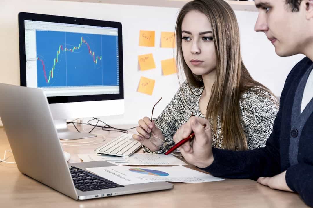 a man and a woman looking at the computer screens with stock graphs